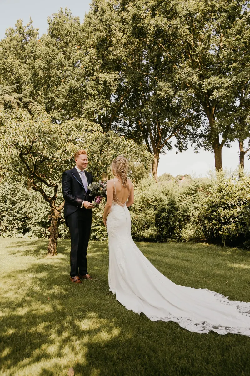 Bruidspaar staat in een tuin, de sleep van de jurk ligt uitgespreid over het gras
