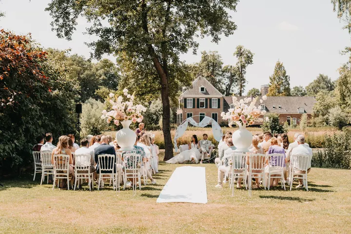Ceremonie in de tuin met gasten op witte stoelen voor een landhuis